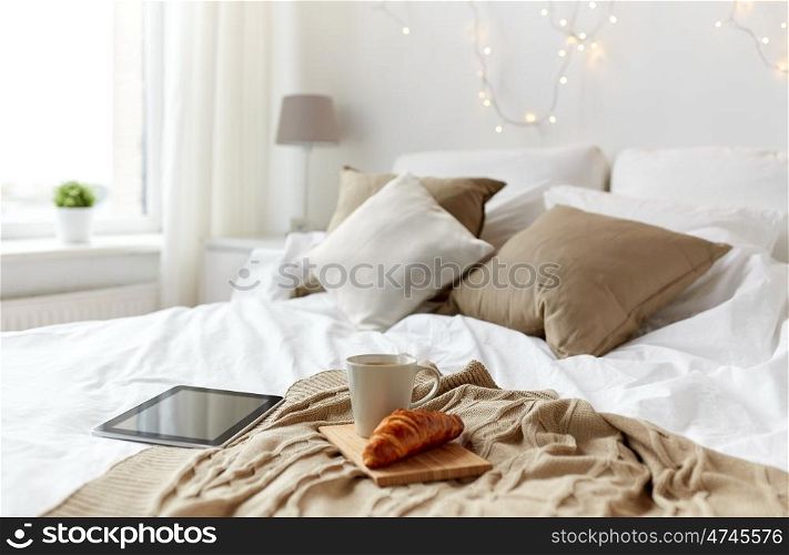 technology, holidays, christmas and winter concept - cozy bedroom with tablet pc computer, coffee cup and croissant on bed at cozy home