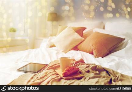 technology, holidays, christmas and winter concept - cozy bedroom with tablet pc computer, coffee cup and croissant on bed at cozy home. tablet pc, coffee cup and croissant on bed at home
