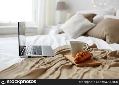 technology, holidays, christmas, interior and winter concept - cozy bedroom with laptop computer, coffee cup and croissant on bed at cozy home