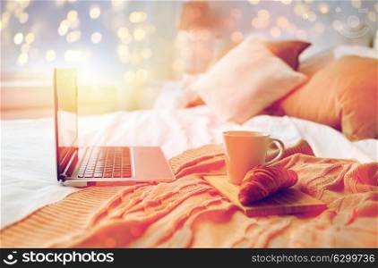 technology, holidays, christmas, interior and winter concept - cozy bedroom with laptop computer, coffee cup and croissant on bed at cozy home. laptop, coffee and croissant on bed at cozy home