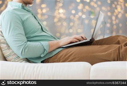 technology, home and lifestyle concept - close up of man working with laptop computer and sitting on sofa over holidays lights background. close up of man working with laptop over lights
