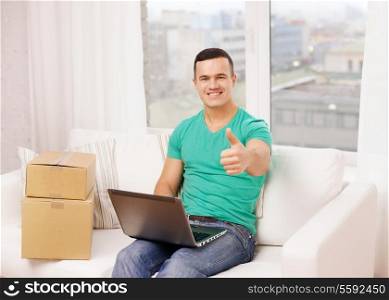 technology, home and lifestyle concept - smiling man with laptop and cardboard boxes at home showing thumbs up