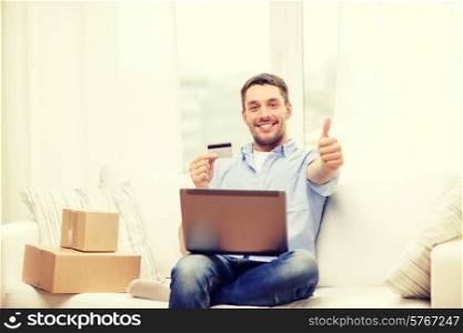 technology, home and lifestyle concept - smiling man with laptop, credit card and cardboard boxes at home showing thumbs up
