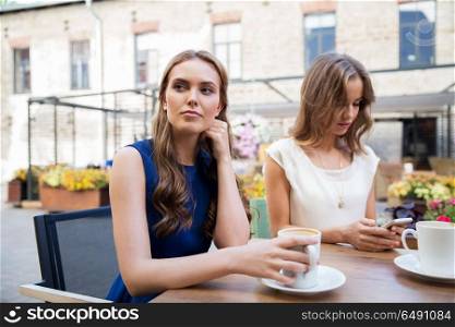 technology, internet addiction and people concept - young woman using smartphone and sad friend drinking coffee at cafe outdoors. young women with smartphone and coffee at cafe. young women with smartphone and coffee at cafe