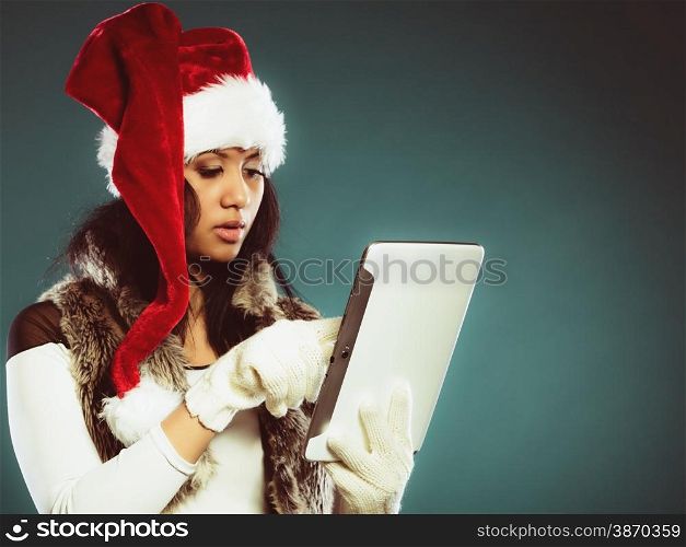 Technology internet and christmas time concept. Mixed race girl in santa claus helper hat using tablet touchpad on blue