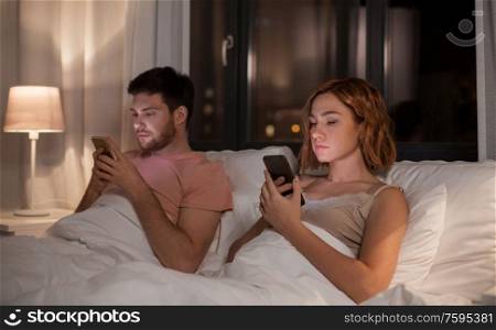 technology, internet and communication concept - unhappy couple using smartphones in bed at night at home. couple using smartphones in bed at night at home