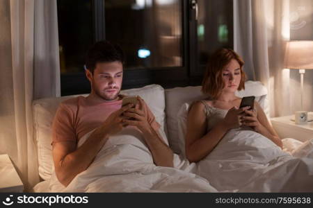 technology, internet and communication concept - unhappy couple using smartphones in bed at night at home. couple using smartphones in bed at night at home