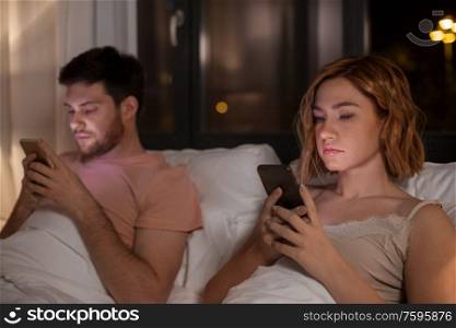 technology, internet and communication concept - unhappy couple using smartphones in bed at night at home. couple using smartphones in bed at night at home