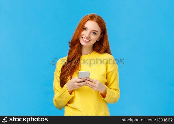 Technology, internet and gadgets concept. Cute redhead woman sending text friend, messaging, having conversation using smartphone app, smiling camera, download useful application, blue background.. Technology, internet and gadgets concept. Cute redhead woman sending text friend, messaging, having conversation using smartphone app, smiling camera, download useful application, blue background