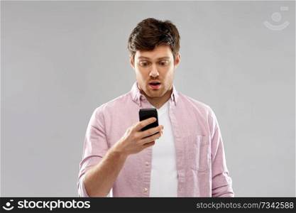 technology, internet and people concept - embarrassed young man looking at something unexpected on smartphone over grey background. embarrassed young man looking at smartphone