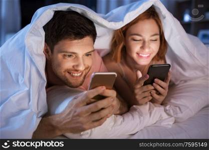 technology, internet and people concept - happy couple using smartphones in bed at night. happy couple using smartphones in bed at night