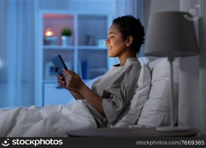 technology, internet and people concept - happy smiling young african american woman with tablet pc computer lying in bed at home at night. woman with tablet pc in bed at home at night