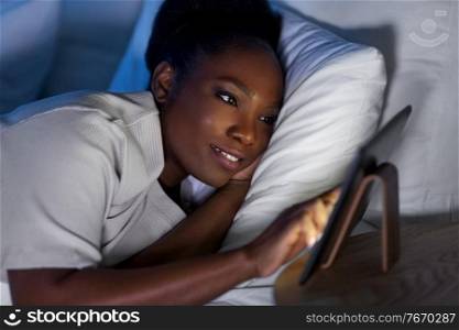 technology, internet and people concept - happy smiling young african american woman with tablet pc computer lying in bed at home at night. woman with tablet pc in bed at home at night