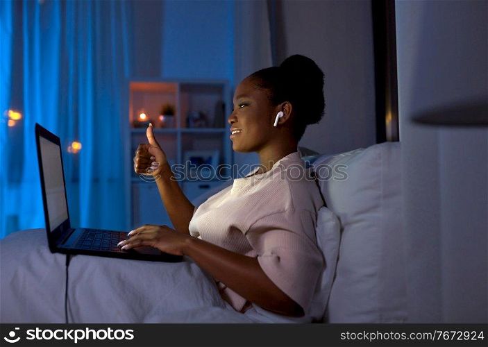 technology, internet and people concept - happy smiling young african american woman with laptop computer and wireless earphones having video call in bed at home at night. woman with laptop having video call at night