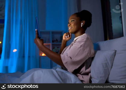 technology, internet and people concept - happy smiling young african american woman with tablet pc computer and wireless earphones lying in bed at home at night. woman with tablet pc in earphones in bed at night