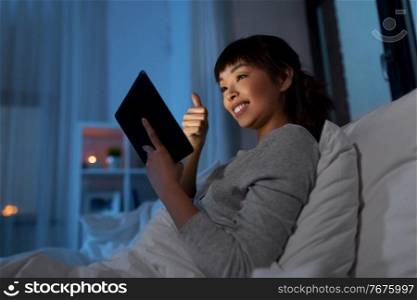 technology, internet and people concept - happy smiling young asian woman in glasses with tablet pc computer having video call in bed at home at night and showing thumbs up. woman with tablet pc in bed has video call at night