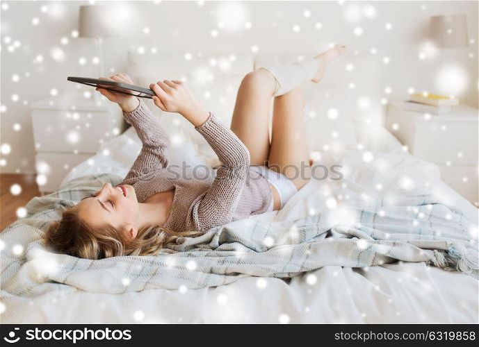 technology, internet and people concept - happy young woman lying in bed with tablet pc computer at home bedroom over snow. happy young woman with tablet pc in bed at home