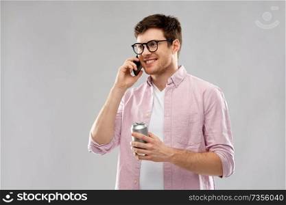 technology, internet and people concept - smiling young man in glasses looking at smartphone and drinking soda from tin can over grey background. young man in glasses with smartphone and drink
