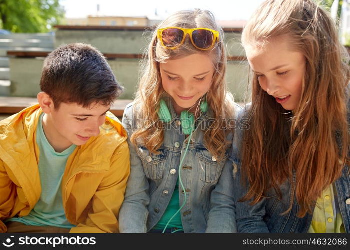 technology, internet and people concept - three happy teenage friends with headphones outdoors looking down at something