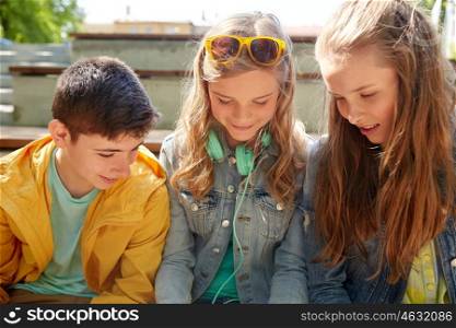 technology, internet and people concept - three happy teenage friends with headphones outdoors looking down at something
