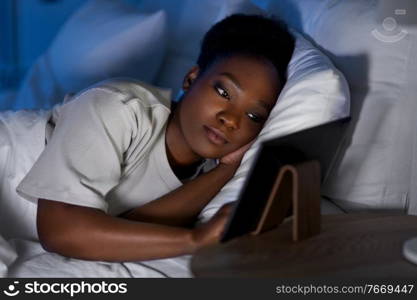 technology, internet and people concept - young african american woman with tablet pc computer lying in bed at home at night. woman with tablet pc in bed at home at night