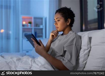 technology, internet and people concept - young african american woman with tablet pc computer lying in bed at home at night. woman with tablet pc in bed at home at night