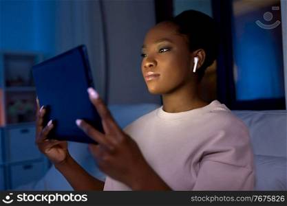 technology, internet and people concept - young african american woman with tablet pc computer and wireless earphones lying in bed at home at night. woman with tablet pc in earphones in bed at night