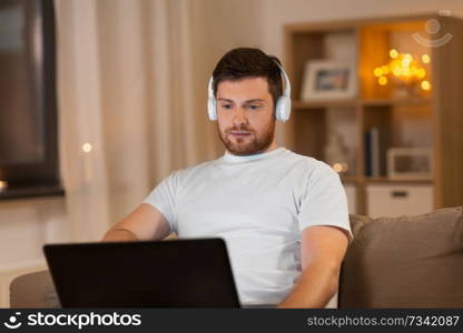 technology, internet and people concept - young man in headphones with laptop computer listening to music at night at home. man in headphones with laptop computer at night