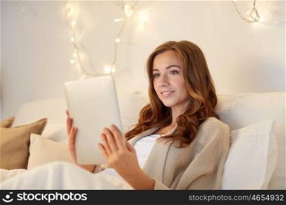technology, internet, christmas and people concept - happy young woman lying in bed with tablet pc computer at home bedroom