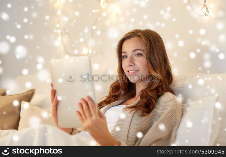 technology, internet, christmas and people concept - happy young woman lying in bed with tablet pc computer at home bedroom over snow. happy young woman with tablet pc in bed at home