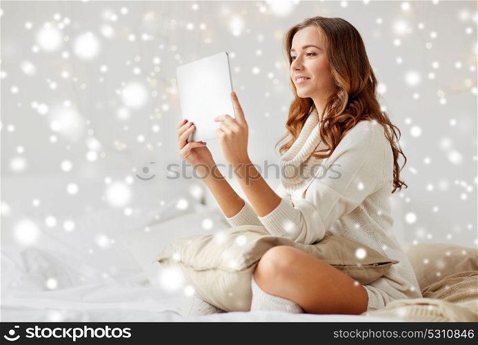 technology, internet, christmas and people concept - happy young woman lying in bed with tablet pc computer at home bedroom over snow. happy young woman with tablet pc in bed at home