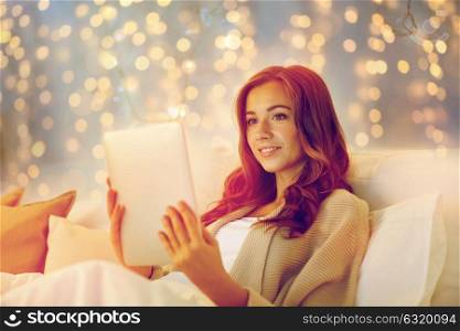 technology, internet, christmas and people concept - happy young woman lying in bed with tablet pc computer at home bedroom. happy young woman with tablet pc in bed at home