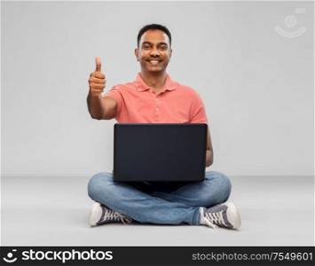 technology, internet, communication and people concept - happy indian man with laptop computer sitting on floor over grey background. happy indian man with laptop showing thumbs up