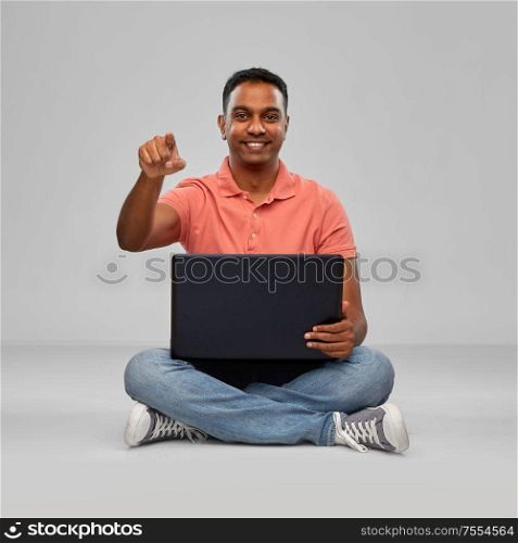technology, internet, communication and people concept - happy indian man with laptop computer sitting on floor and pointing fnger to camera over grey background. happy indian man with laptop computer
