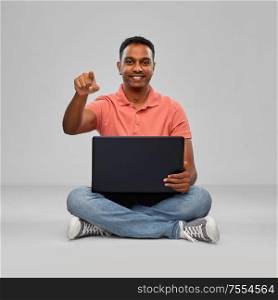 technology, internet, communication and people concept - happy indian man with laptop computer sitting on floor and pointing fnger to camera over grey background. happy indian man with laptop computer