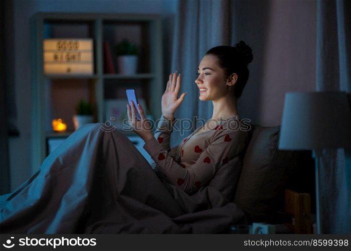 technology, internet, communication and people concept - happy smiling teenage girl with smartphone having video call sitting in bed at home at night. teenage girl with phone having video call in bed