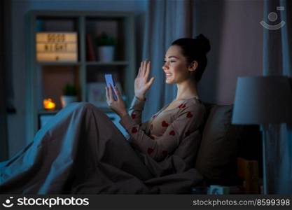 technology, internet, communication and people concept - happy smiling teenage girl with smartphone having video call sitting in bed at home at night. teenage girl with phone having video call in bed