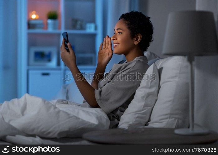 technology, internet, communication and people concept - happy smiling young african american woman with smartphone having video call lying in bed at home at night. woman with phone having video call in bed at night
