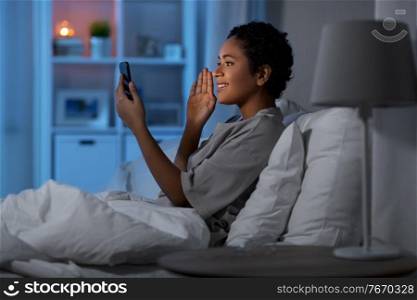 technology, internet, communication and people concept - happy smiling young african american woman with smartphone having video call lying in bed at home at night. woman with phone having video call in bed at night