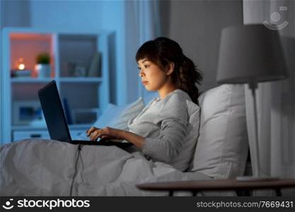 technology, internet, communication and people concept - happy smiling young asian woman with laptop computer lying in bed at home at night. woman with laptop in bed at home at night