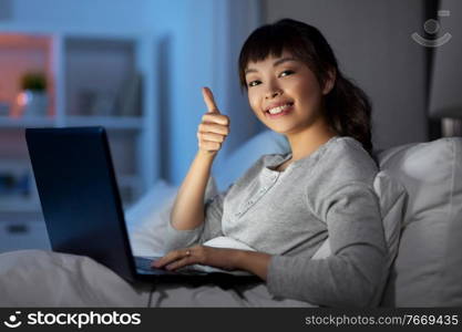 technology, internet, communication and people concept - happy smiling young asian woman with laptop computer lying in bed at home at night and showing thumbs up. woman with laptop in bed at night shows thumbs up