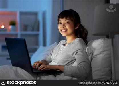 technology, internet, communication and people concept - happy smiling young asian woman with laptop computer lying in bed at home at night. woman with laptop in bed at home at night