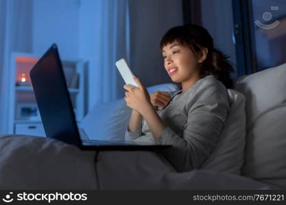 technology, internet, communication and people concept - happy smiling young asian woman with smartphone and laptop lying in bed at home at night. asian woman with smartphone in bed at night