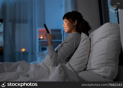 technology, internet, communication and people concept - happy smiling young asian woman with smartphone lying in bed at home at night. asian woman with smartphone in bed at night