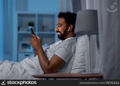 technology, internet, communication and people concept - happy smiling young indian man with smartphone and earphones listening to music in bed at night. man with smartphone and earphones in bed at night