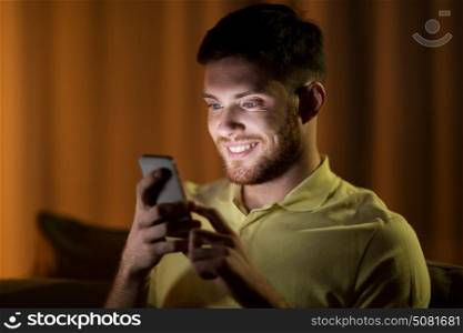 technology, internet, communication and people concept - happy smiling young man texting on smartphone at home at night. happy young man with smartphone at night