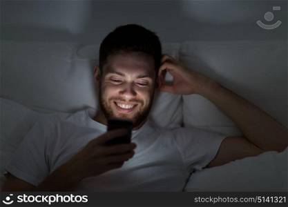 technology, internet, communication and people concept - happy smiling young man texting on smartphone in bed at home at night. happy young man with smartphone in bed at night