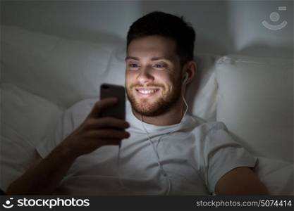 technology, internet, communication and people concept - happy smiling young man with smartphone and earphones listening to music in bed at night. man with smartphone and earphones in bed at night