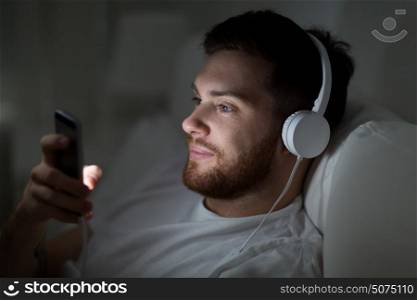technology, internet, communication and people concept - happy smiling young man with smartphone and headphones hones listening to music in bed at night. man with smartphone and headphones in bed at night