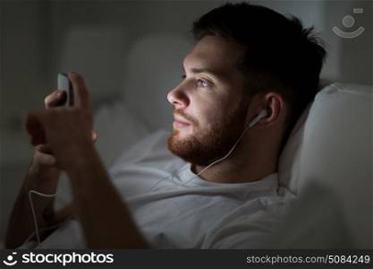 technology, internet, communication and people concept - happy smiling young man with smartphone and earphones listening to music in bed at night. man with smartphone and earphones in bed at night. man with smartphone and earphones in bed at night